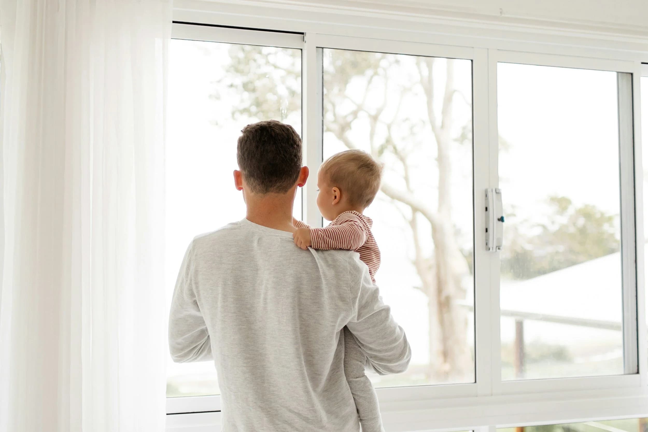 a man with his back to the camera looking out of a bright window whilst holding a toddler