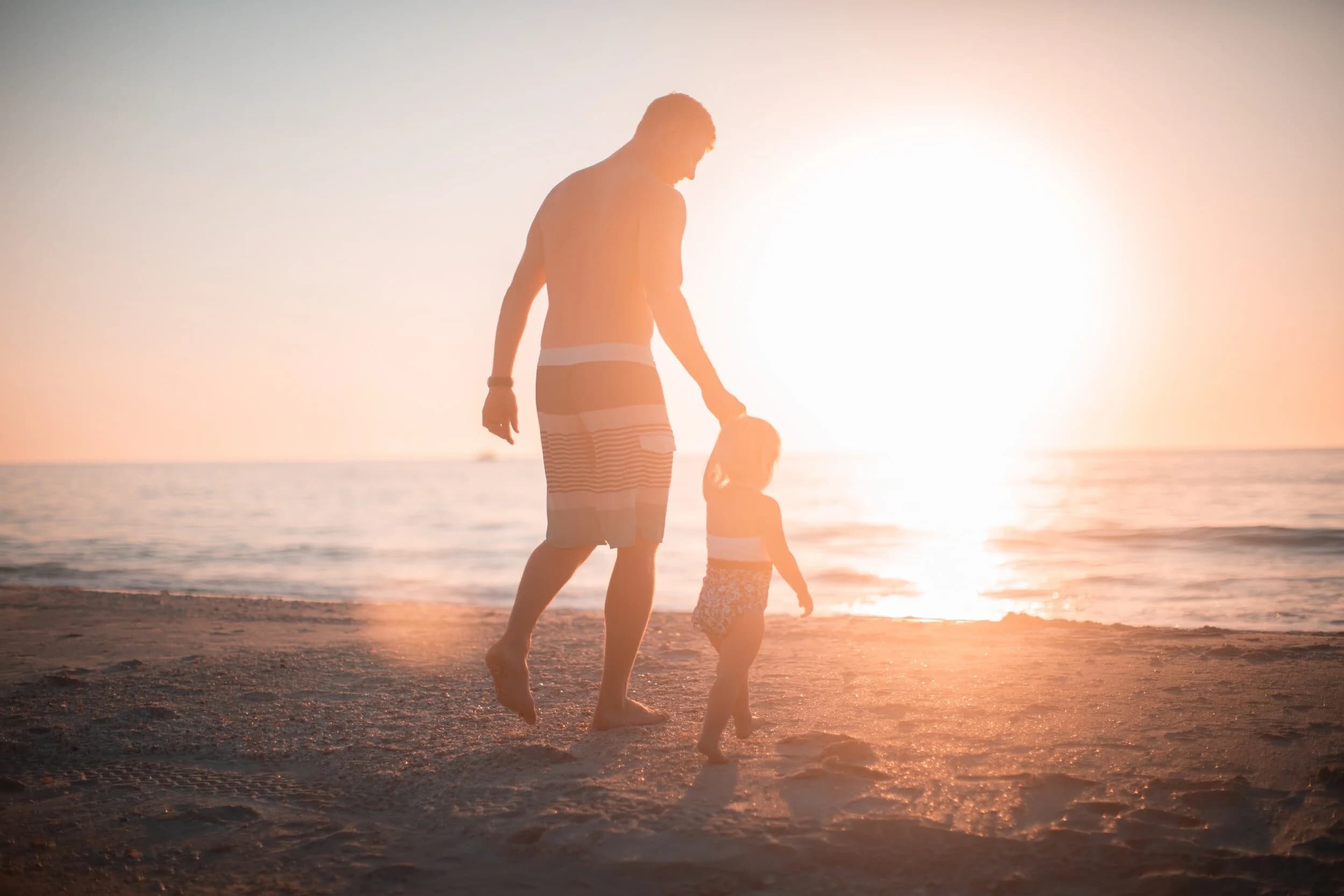 A man walks hand in hand with a small child on a beach. The sun is shining.